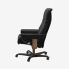 A black leather high-back office chair with a footrest, featuring a dark wood central column and a five-star black metal base with casters, shown from a side profile on a light gray background.